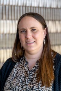 **Alt text:** Portrait of a woman with long brown hair wearing a patterned blouse and black cardigan, standing in front of a neutral textured background.