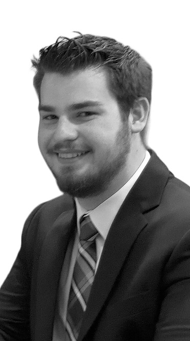 Black-and-white portrait of a smiling man with short hair and a beard wearing a suit and striped tie, seated at an event.