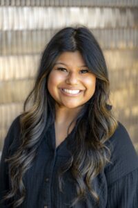 Portrait of a smiling woman with long dark hair wearing a black blouse, standing in front of a neutral textured background.