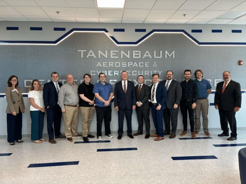 A group of thirteen people standing in a line and smiling inside the Tanenbaum Aerospace & Cybersecurity Center at Rose State College, posing in front of a wall with the center’s name displayed.