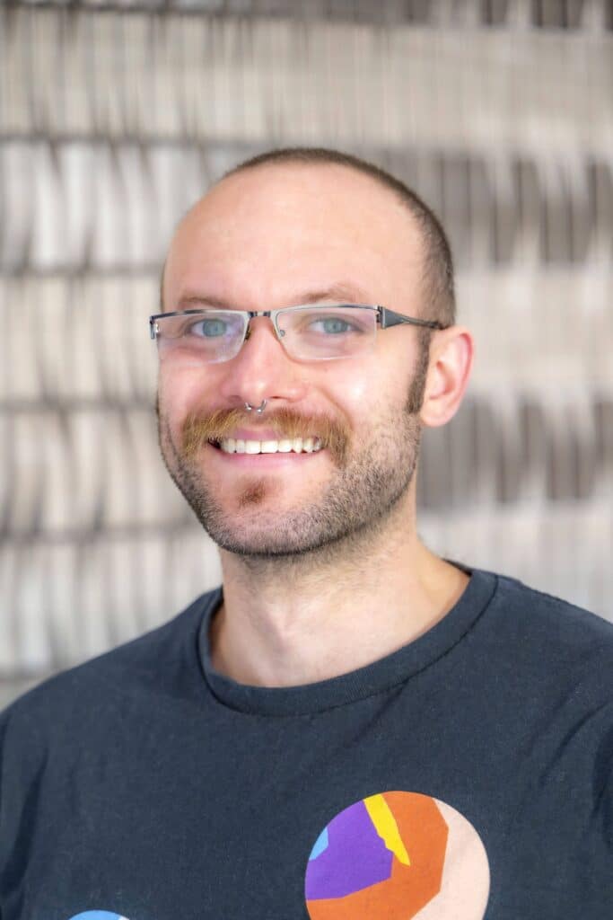 Professional headshot of a smiling man with glasses, short hair, and a mustache, wearing a dark T-shirt with a colorful circular design, in front of a softly blurred neutral background.