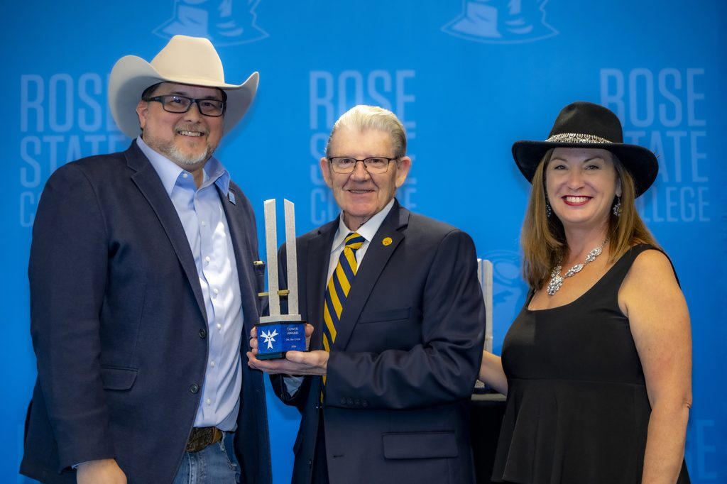 Dr. Jim Cook, center, holds his Rose State College Tower Award while posing with two college representatives in cowboy hats against a blue Rose State College backdrop.