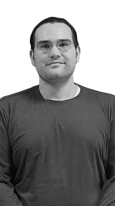 Black-and-white portrait of a man with glasses and short dark hair wearing a long-sleeve shirt, standing and looking at the camera.