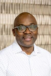 Professional headshot of a smiling man wearing glasses and a white dress shirt with a subtle pattern, in front of a softly blurred neutral background.