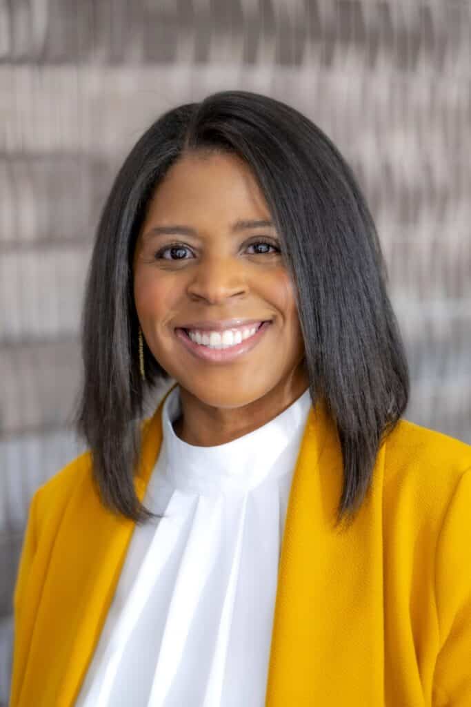 Professional headshot of a smiling woman with straight shoulder-length dark hair, wearing a white blouse and mustard yellow blazer, in front of a softly blurred neutral background.