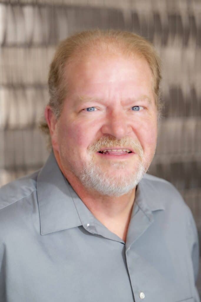 Professional headshot of a smiling man with short light hair and a trimmed beard, wearing a gray collared shirt, in front of a softly blurred neutral background.