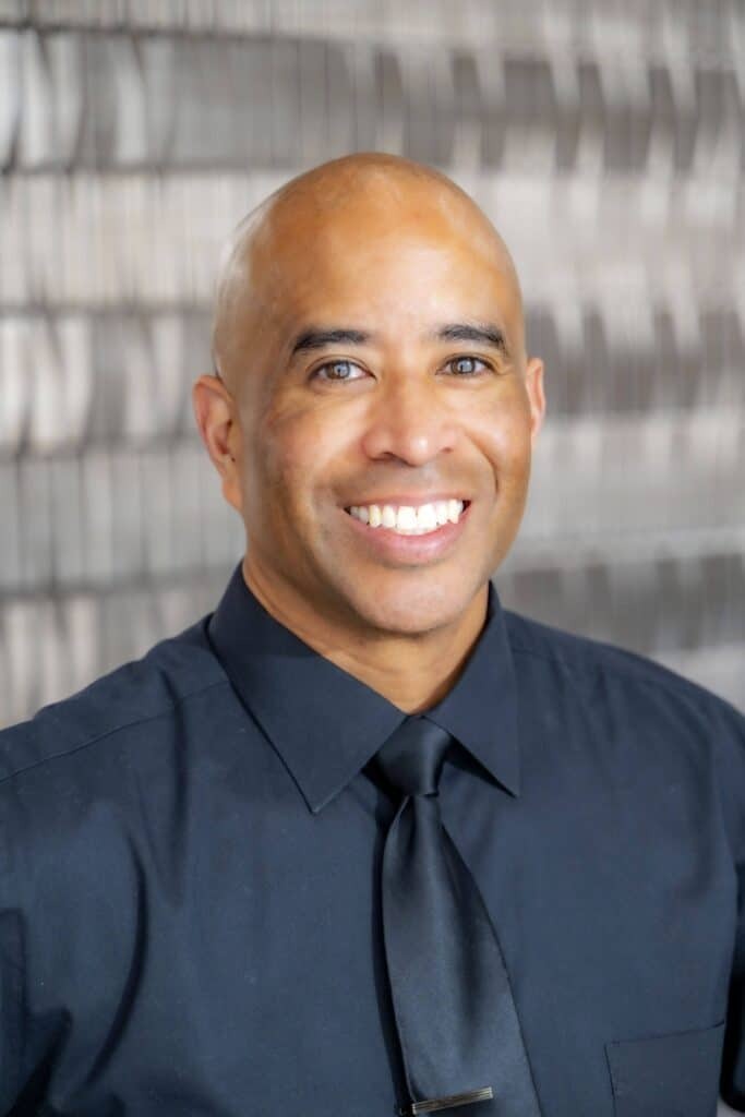 Professional headshot of a smiling man wearing a black dress shirt and black tie, in front of a softly blurred neutral background.