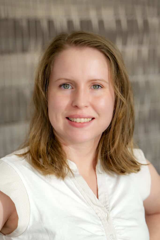 Professional headshot of a smiling woman with shoulder-length light brown hair, wearing a white sleeveless blouse, in front of a softly blurred neutral background.