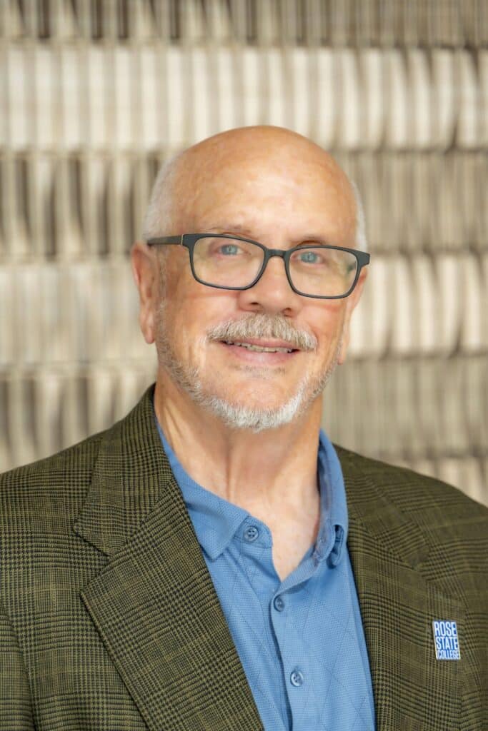 Professional headshot of a smiling man with glasses and a short gray beard wearing a blue shirt and green plaid blazer with a Rose State College pin, in front of a softly blurred neutral background.