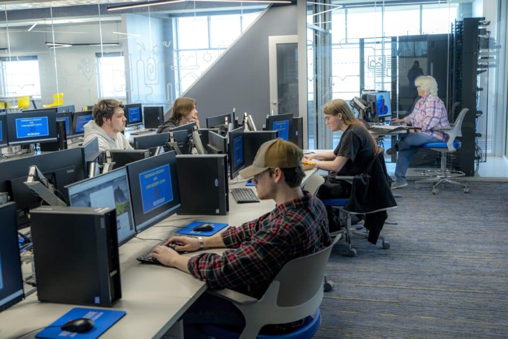 Students work at desktop computers in a modern cybersecurity classroom at Rose State College. Several students focus on coding and cybersecurity exercises while an instructor assists in the background. The classroom features glass walls, bright lighting, and multiple computer stations.