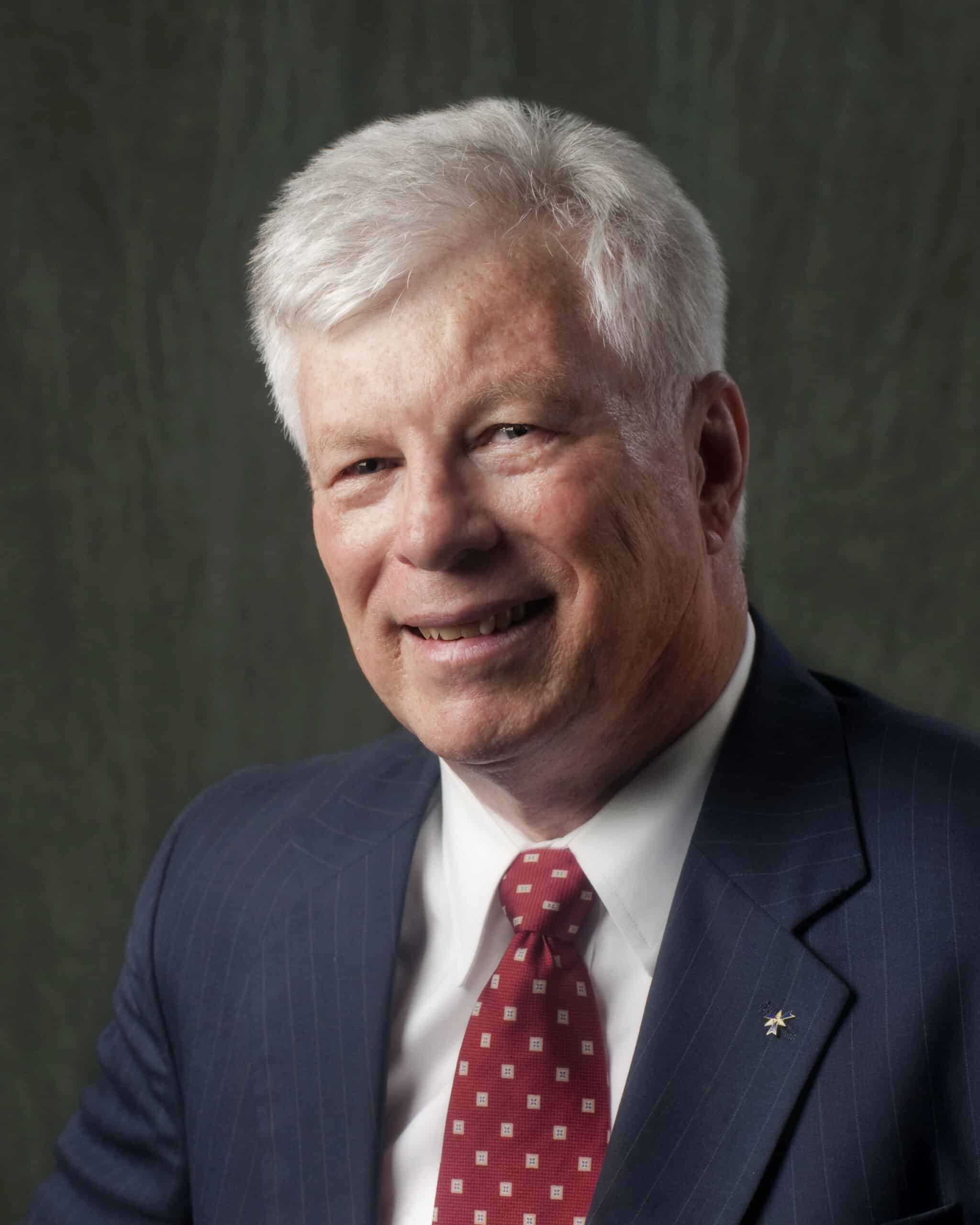 Professional headshot of a smiling man with short white hair wearing a dark pinstripe suit, white dress shirt, and red patterned tie, posed in front of a plain dark background.