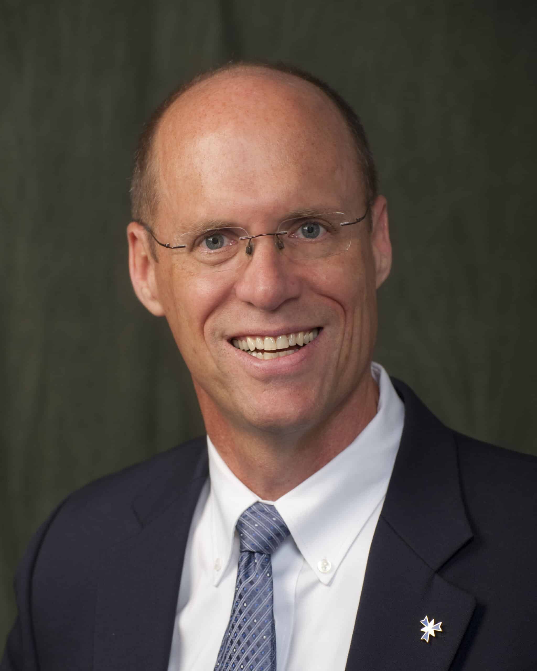Professional headshot of a smiling man with glasses wearing a dark suit jacket, white dress shirt, and light blue patterned tie, posed in front of a plain dark background.