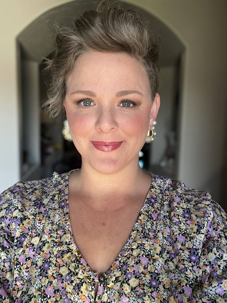 Professional headshot of a smiling woman with short wavy hair wearing pearl earrings and a floral blouse with shades of purple, yellow, and white, in a softly lit indoor setting.