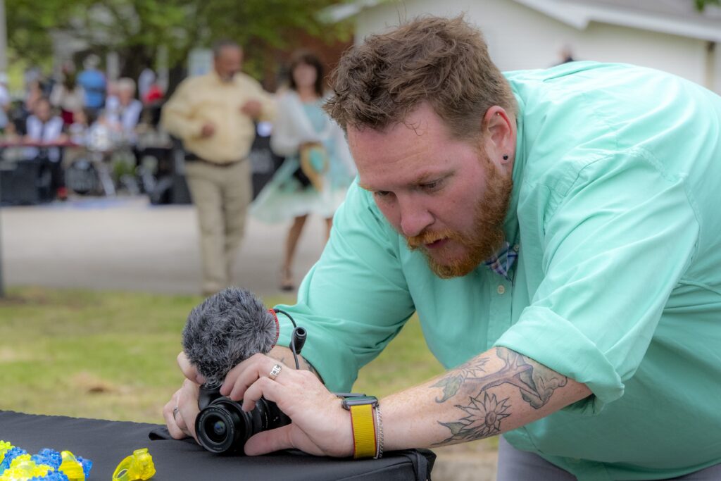 Man in a teal shirt adjusting a camera on a table outdoors, with people and event activity blurred in the background.