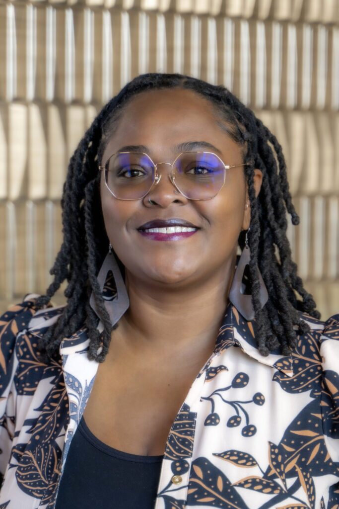 Smiling woman with glasses wearing large earrings and a patterned shirt against a softly blurred neutral background.
