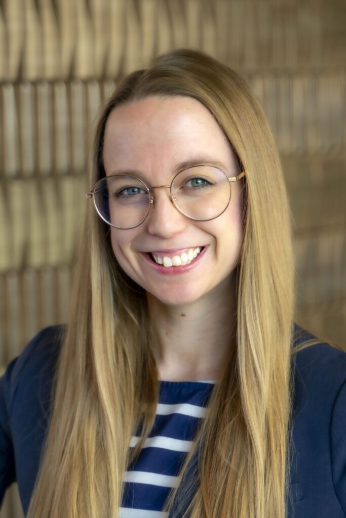 Smiling woman with long straight blonde hair and round glasses, wearing a navy blazer over a navy-and-white striped top.