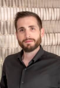 Professional headshot of a man with short brown hair and a trimmed beard wearing a black button-up shirt, in front of a softly blurred neutral background.