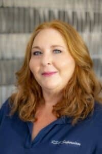 Professional headshot of a smiling woman with shoulder-length auburn hair, wearing a navy blue shirt embroidered with 'RSC Supervisor,' in front of a softly blurred neutral background.