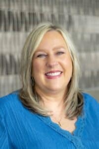 Professional headshot of a smiling woman with blonde hair wearing a blue blouse, in front of a softly blurred neutral background.