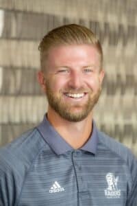 Professional headshot of a smiling man with short light brown hair and beard wearing a gray Adidas Rose State Raiders polo shirt, in front of a softly blurred neutral background.