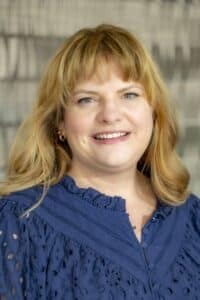 Professional headshot of a smiling woman with blonde hair wearing a navy blue blouse with eyelet details, in front of a softly blurred neutral background.