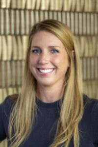 Smiling woman with long blonde hair wearing a dark top against a softly blurred neutral background.