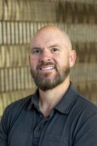 Smiling bald man with a full beard wearing a dark gray collared shirt.