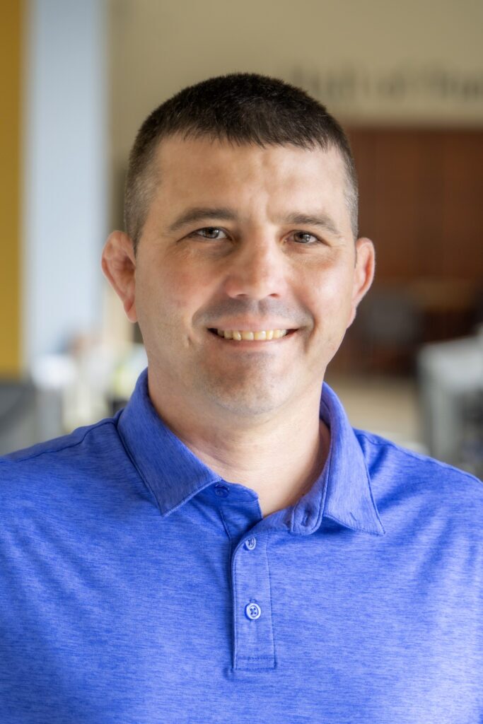Portrait of a smiling man wearing a blue collared shirt, standing indoors with a softly blurred background.