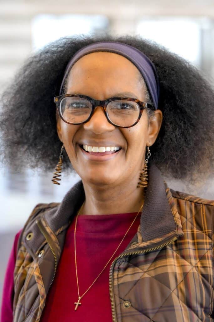 Professional headshot of a smiling woman wearing glasses, a purple headband, brown plaid vest, and red top with a cross necklace, posed in front of a softly blurred background.